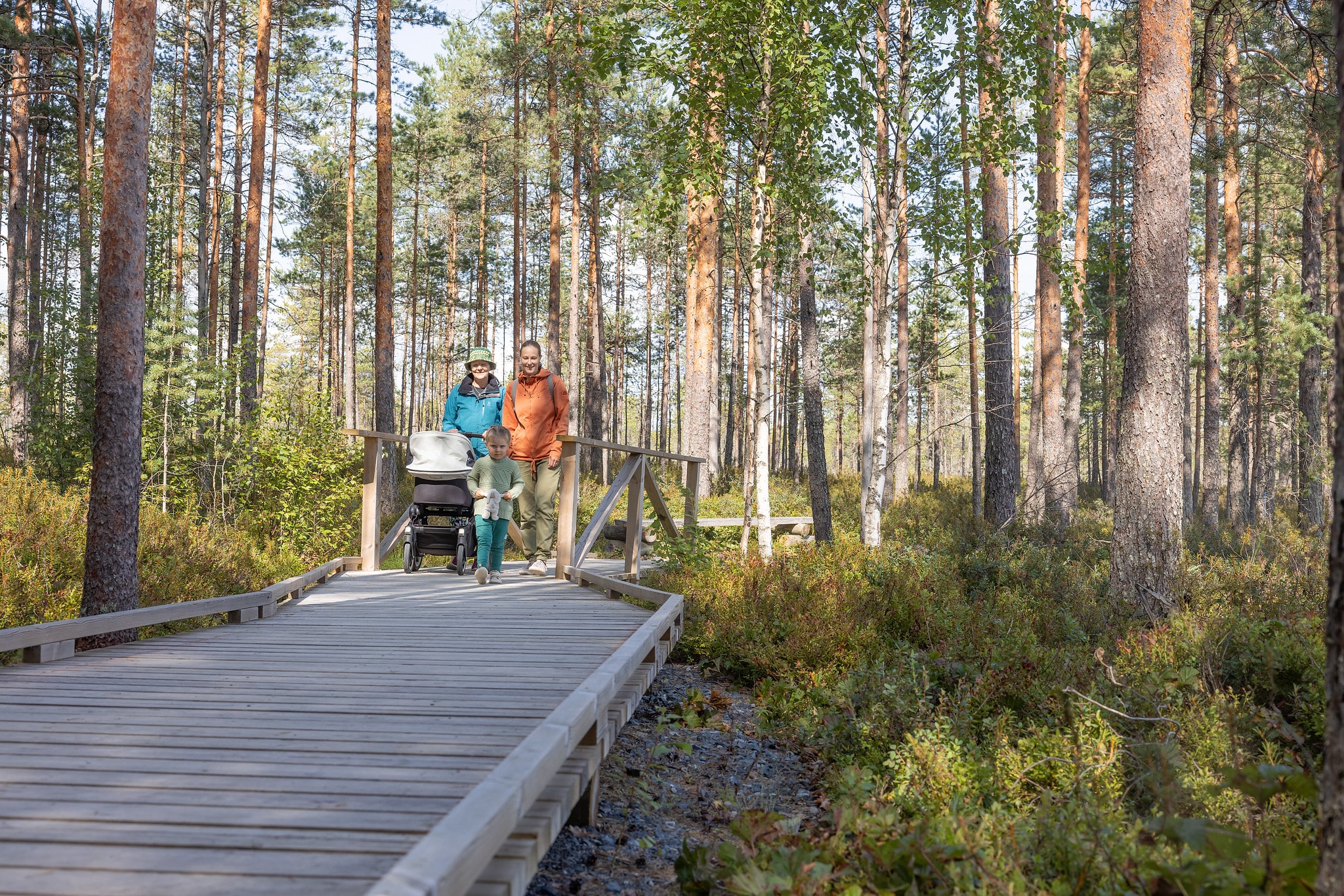 Kaksi naista kävelee puusta rakennetulla leveällä reitillä metsässä. Toinen nainen työntää lastenvaunuja. Edessä juoksee pieni lapsi.