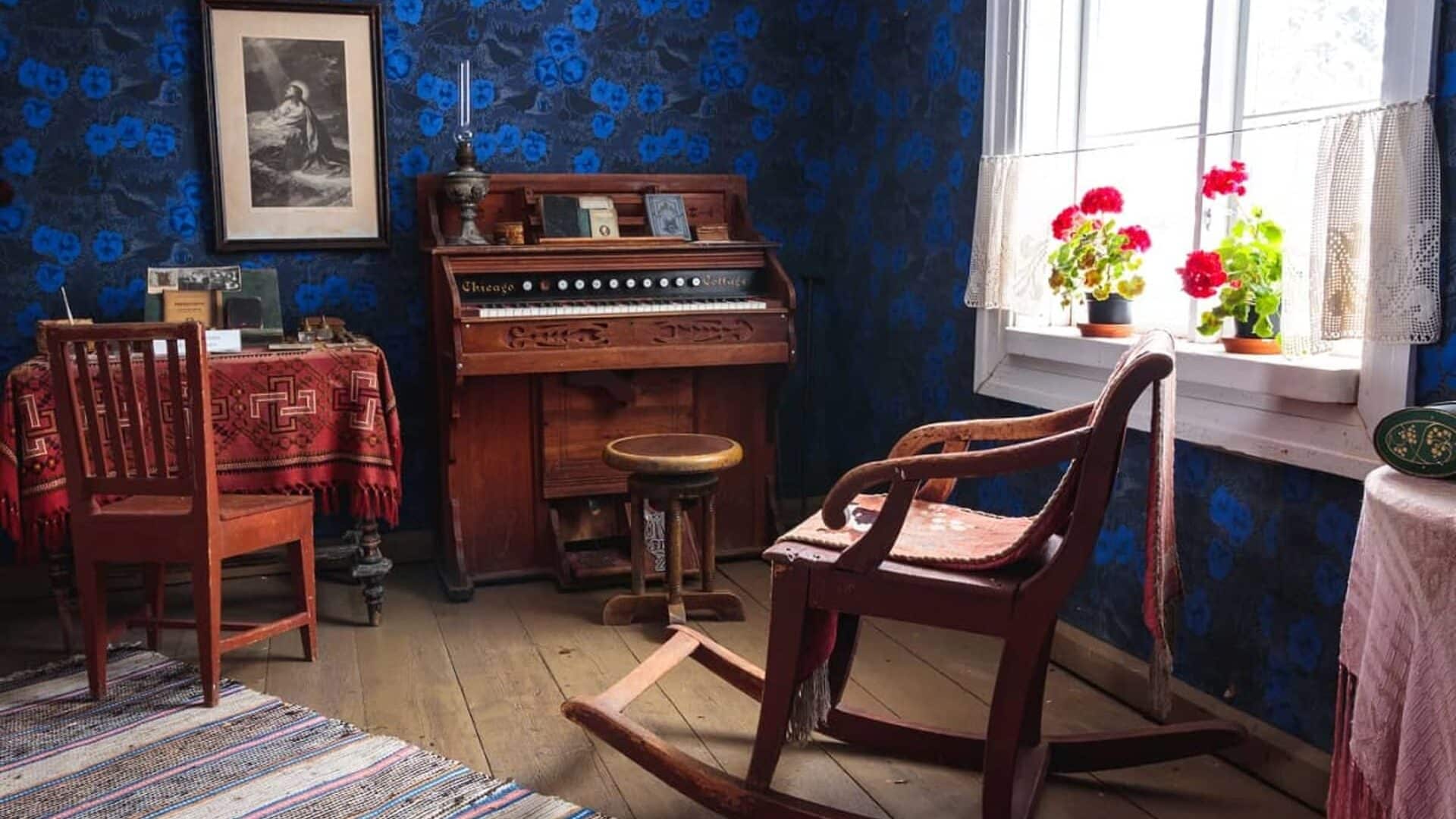 Inside the old house is an old harmonium with a low round chair in front of it. Next to the harmonium is a window with red geraniums and an old wooden rocking chair.