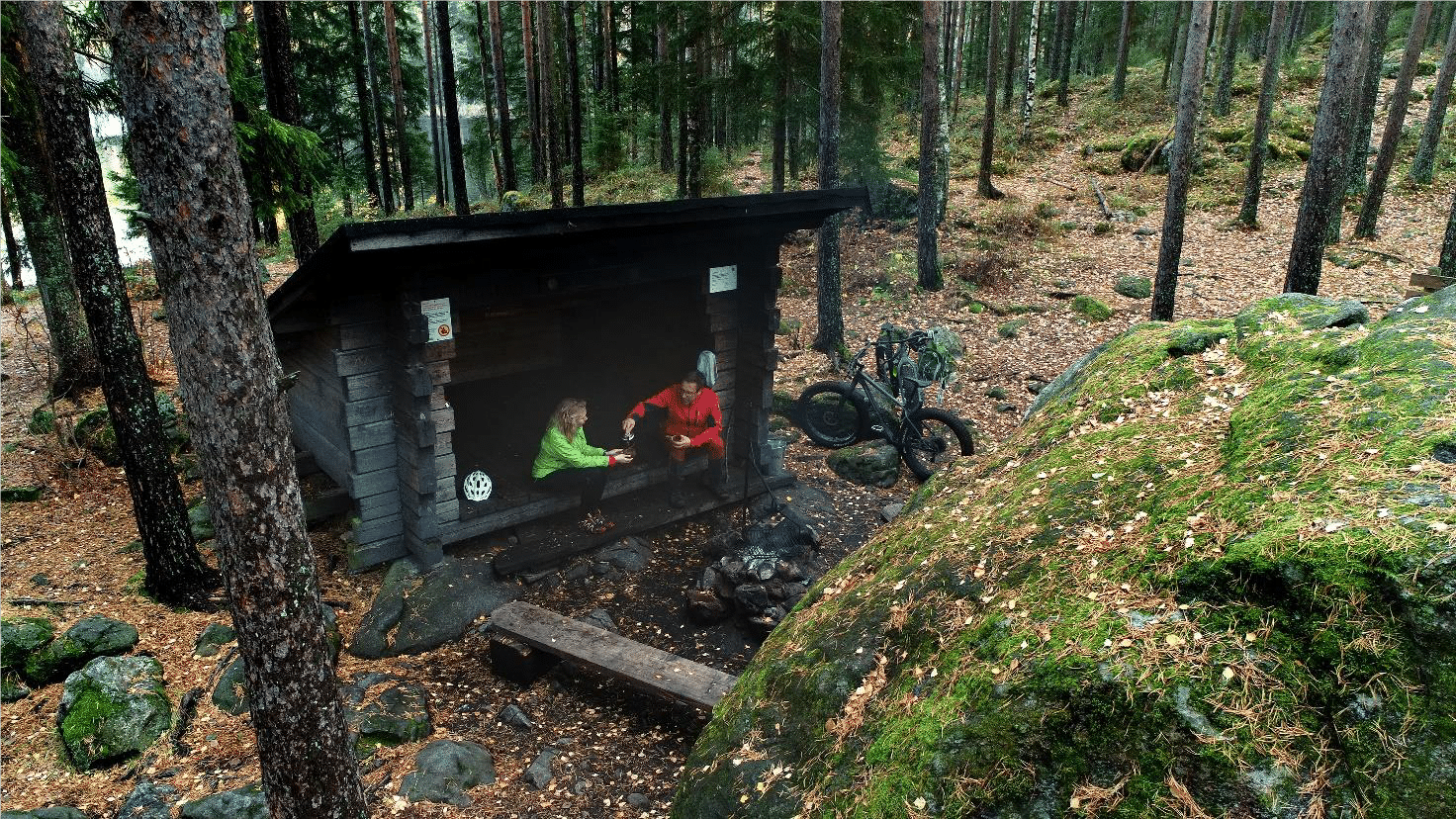 Kaksi henkilöä ovat laavulla. Toinen kaataa lämmintä juomaa toisen kuksaan. Vieressä on maastopyörä.