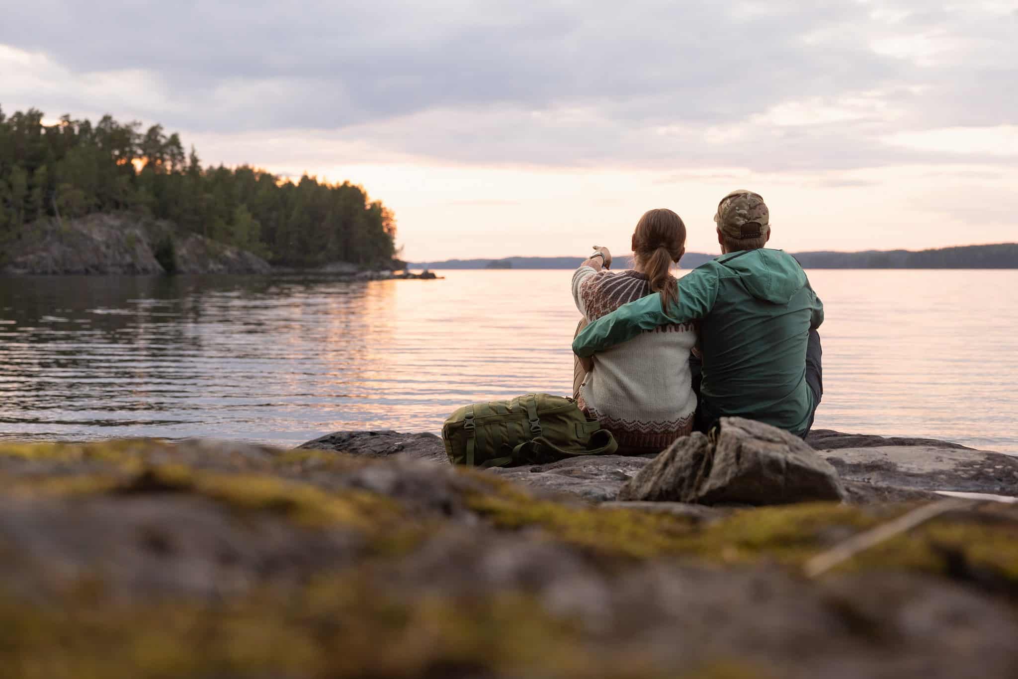 A woman and a man are sitting on a rock, watching the sunset over the lake.