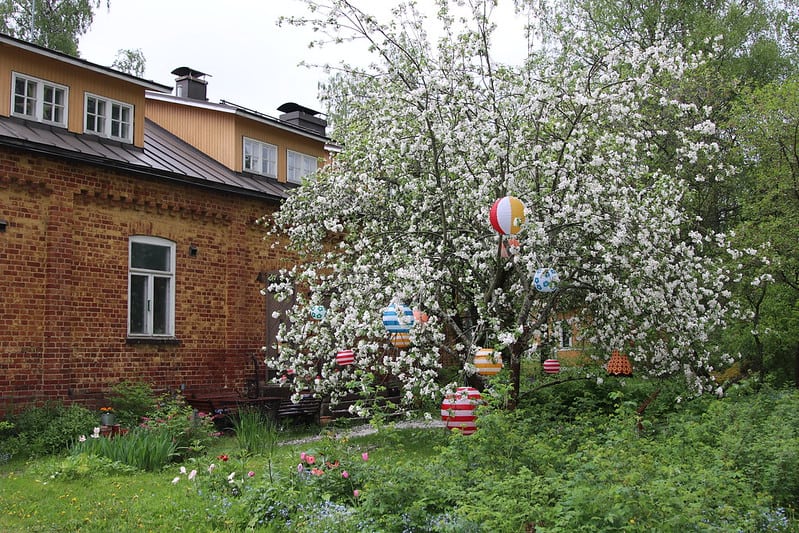 Omenapuu kukkii valkoisena. Puuhun on kiinnitetty värikkäitä koristeita. Taustalla on punatiilinen rakennus.