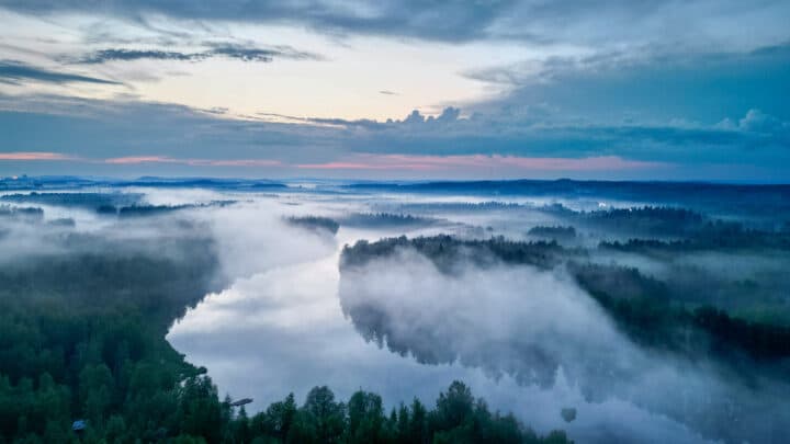 Sunset paints the sky pink and blue. A fog is setting down on a lake and river nestled between a forest.