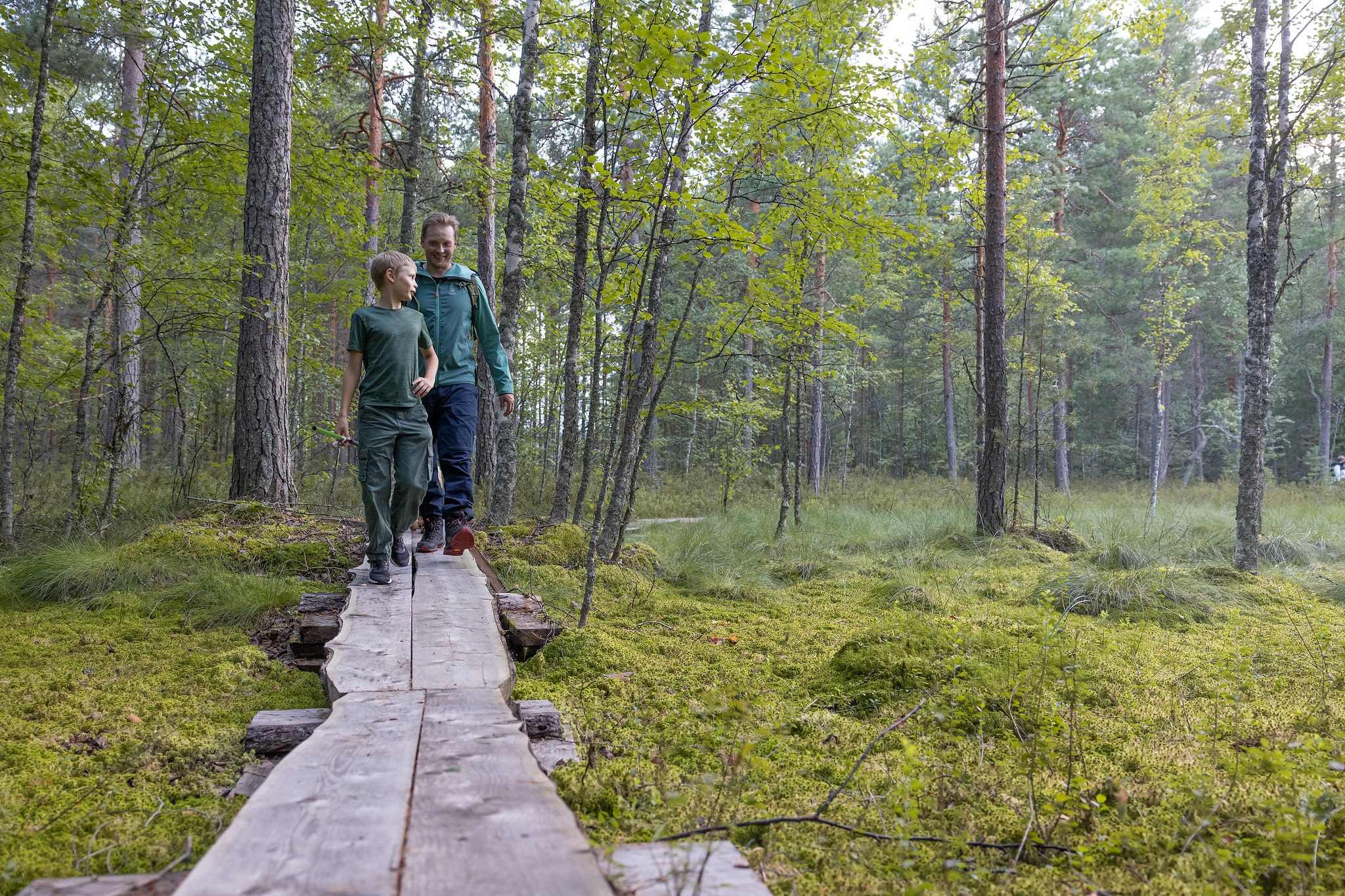 Poika ja mies kävelevät pitkospuilla. Poika juttelee ja katsoo taakseen. Mies hymyilee hänelle.