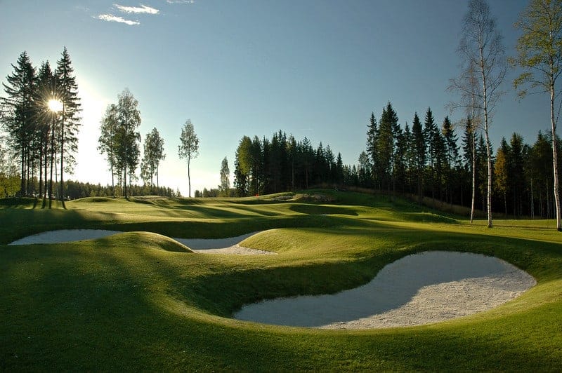 Linnagolf golfkenttä vihreää nurmea ja hiekkakuoppia, taustalla puita joiden välistä paistaa aurinko.