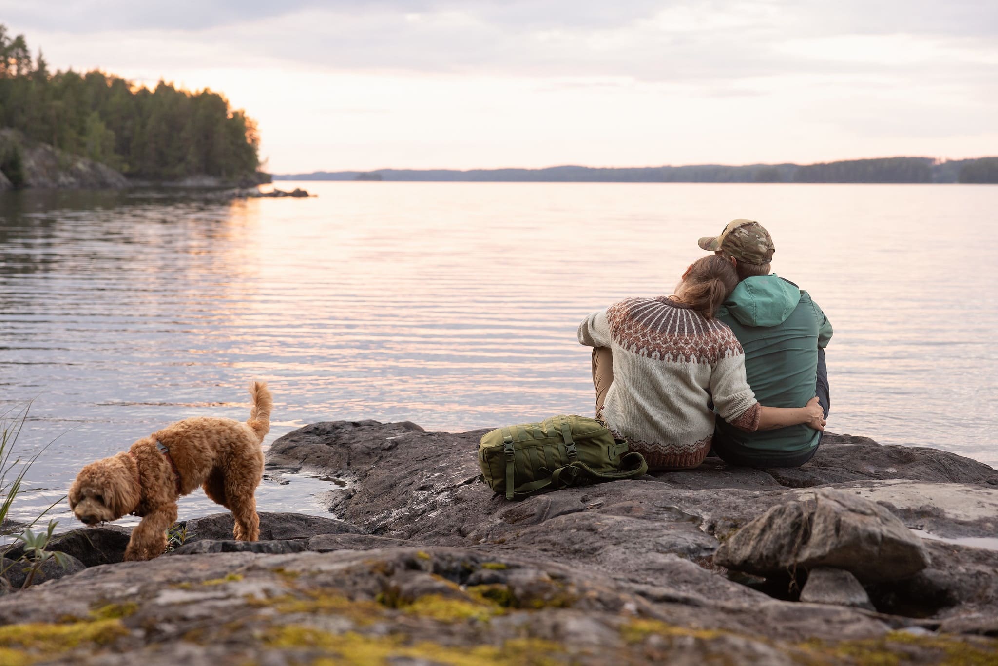 Man and woman sitting at the rock beside the lake with their dog wathcing sunset.