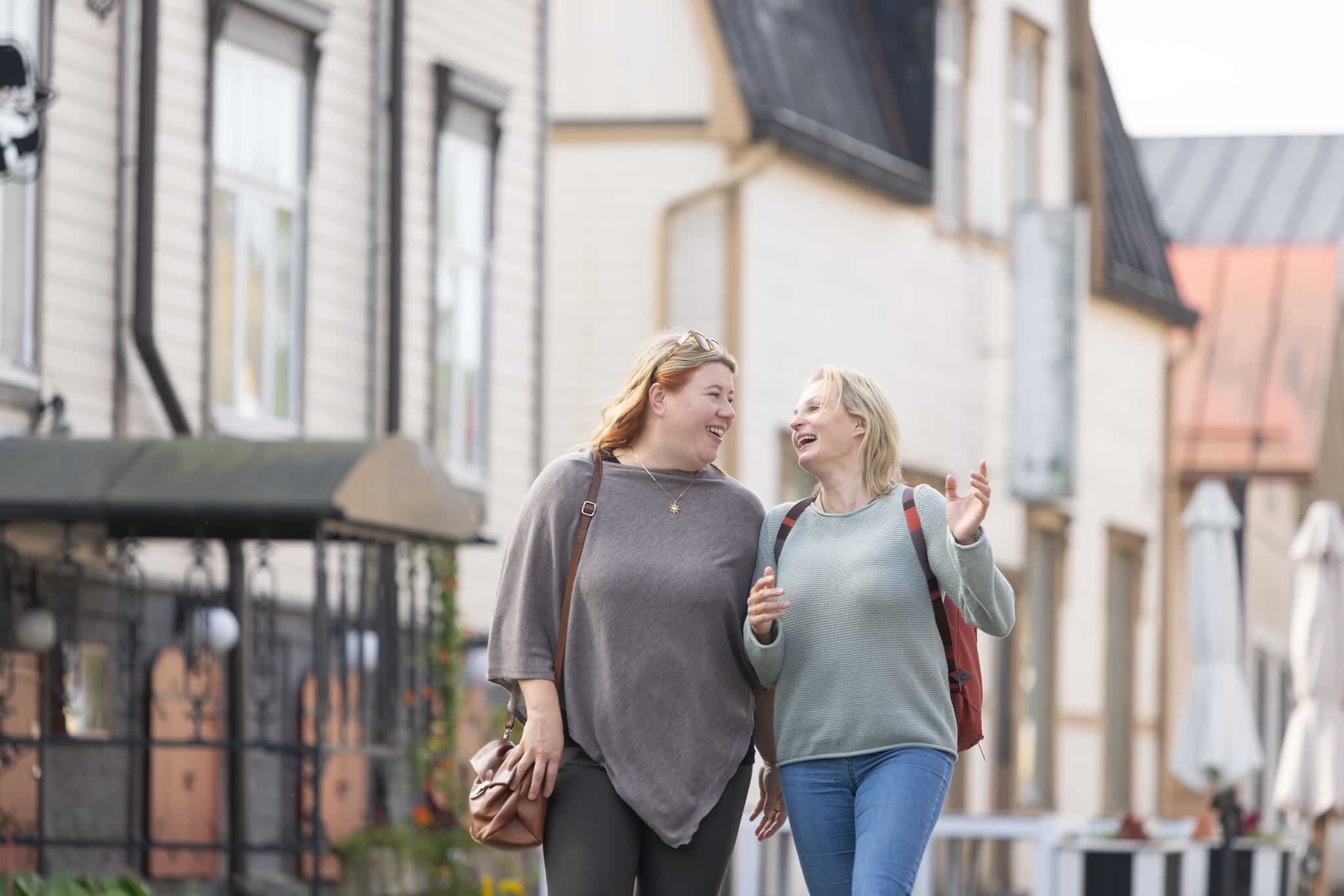 Two women walking and looking at each other smiling, in front of old buildings in the city.