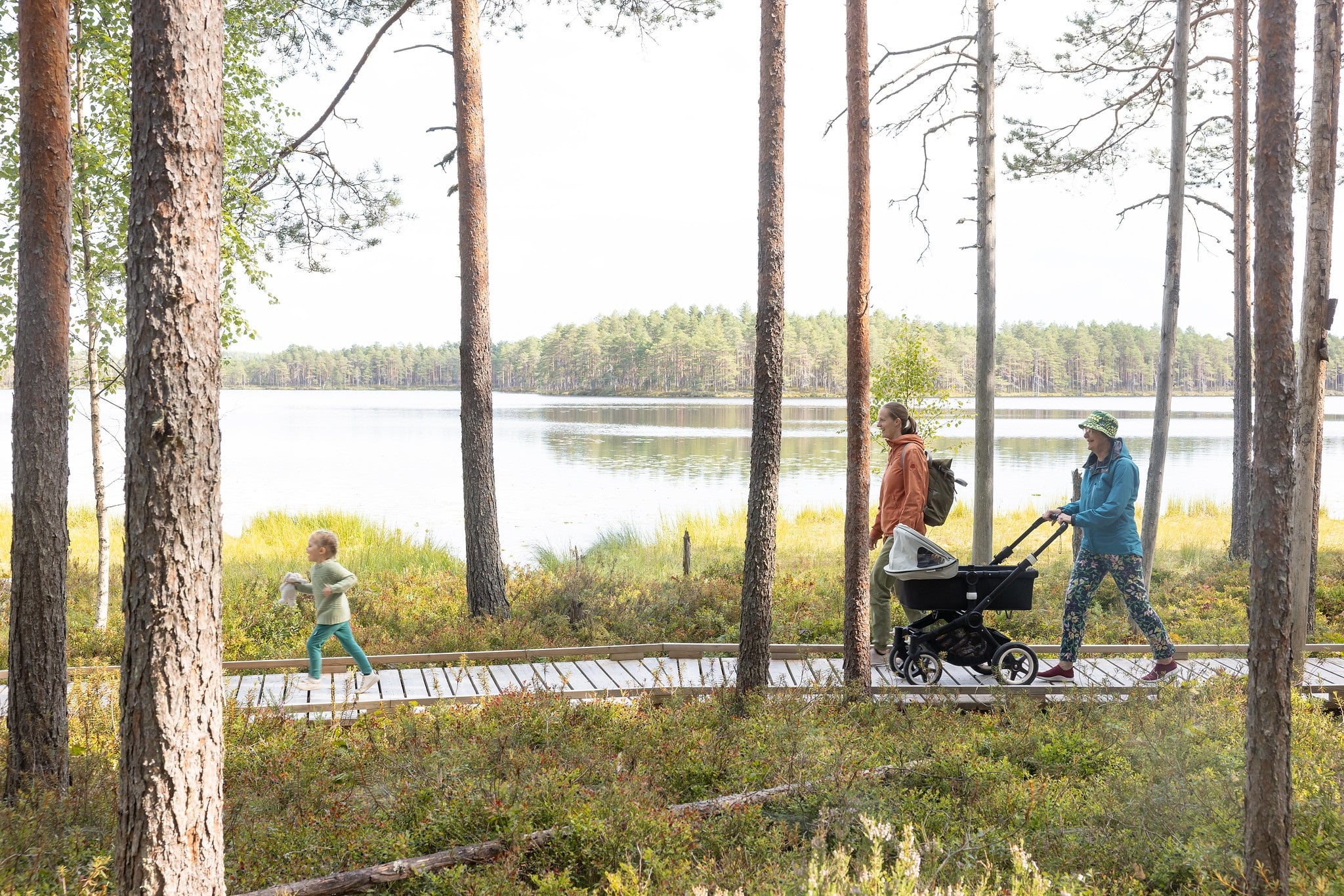 Kaksi naista ja lapsi sekä lastenvaunut pitkospuilla metsässä ja taustalla järvimaisema