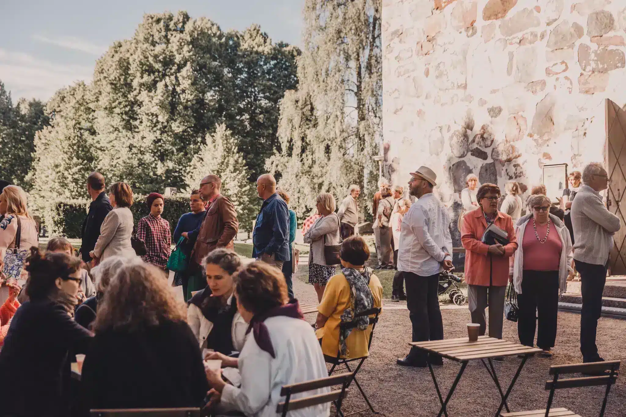 People have gathered in front of the medieval stone church. They talk to each other.