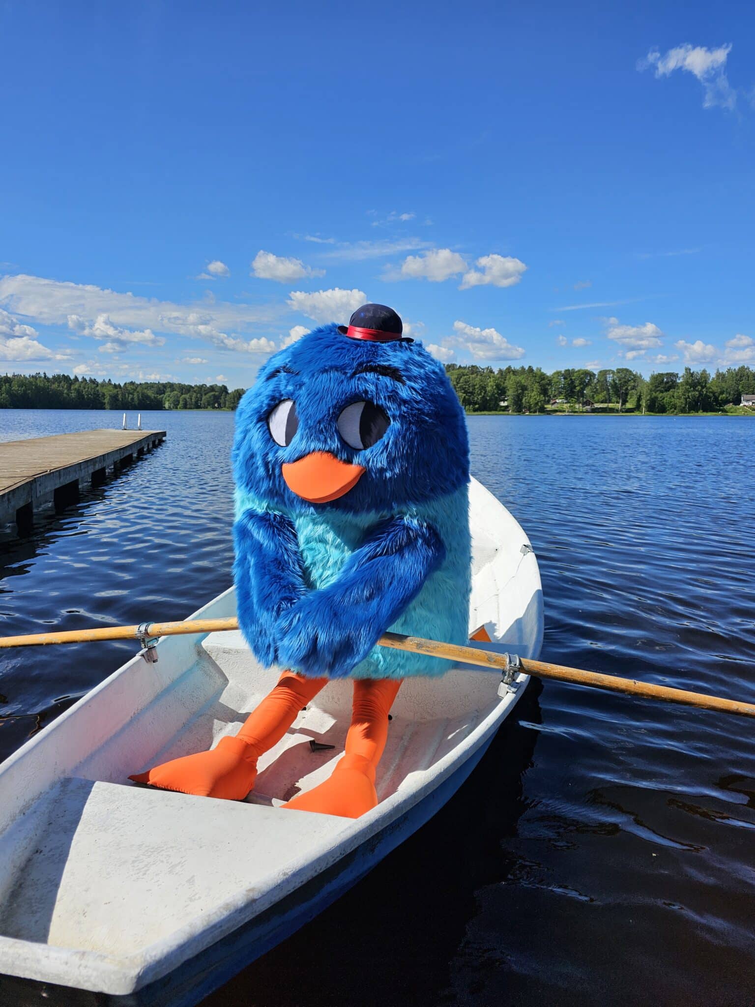 Sininen lintumaskotti istuu soutuveneessä järvellä airot kädessä.