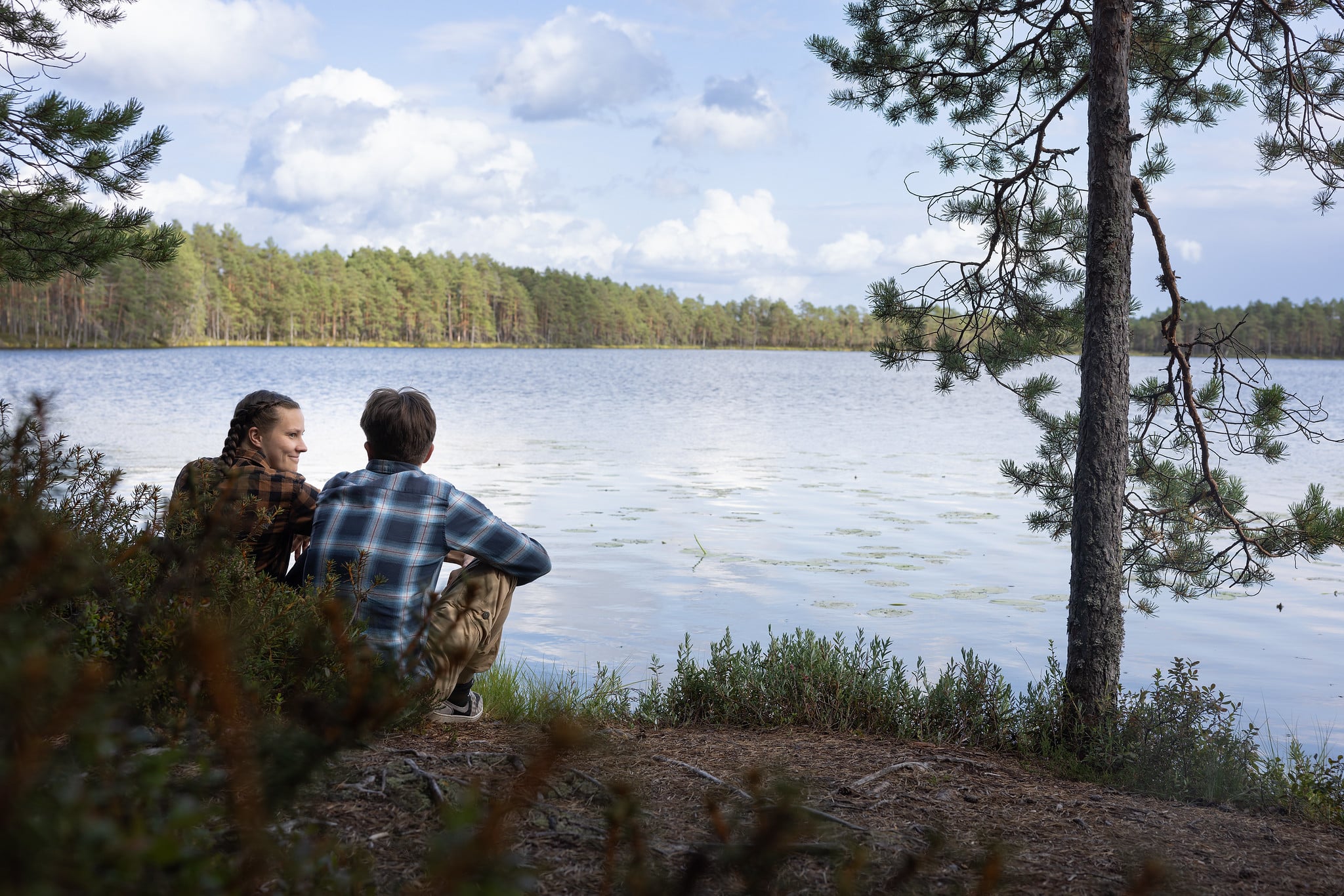 Kaksi ihmistä istuvat järven rannalla. Taustalla näkyy metsää.