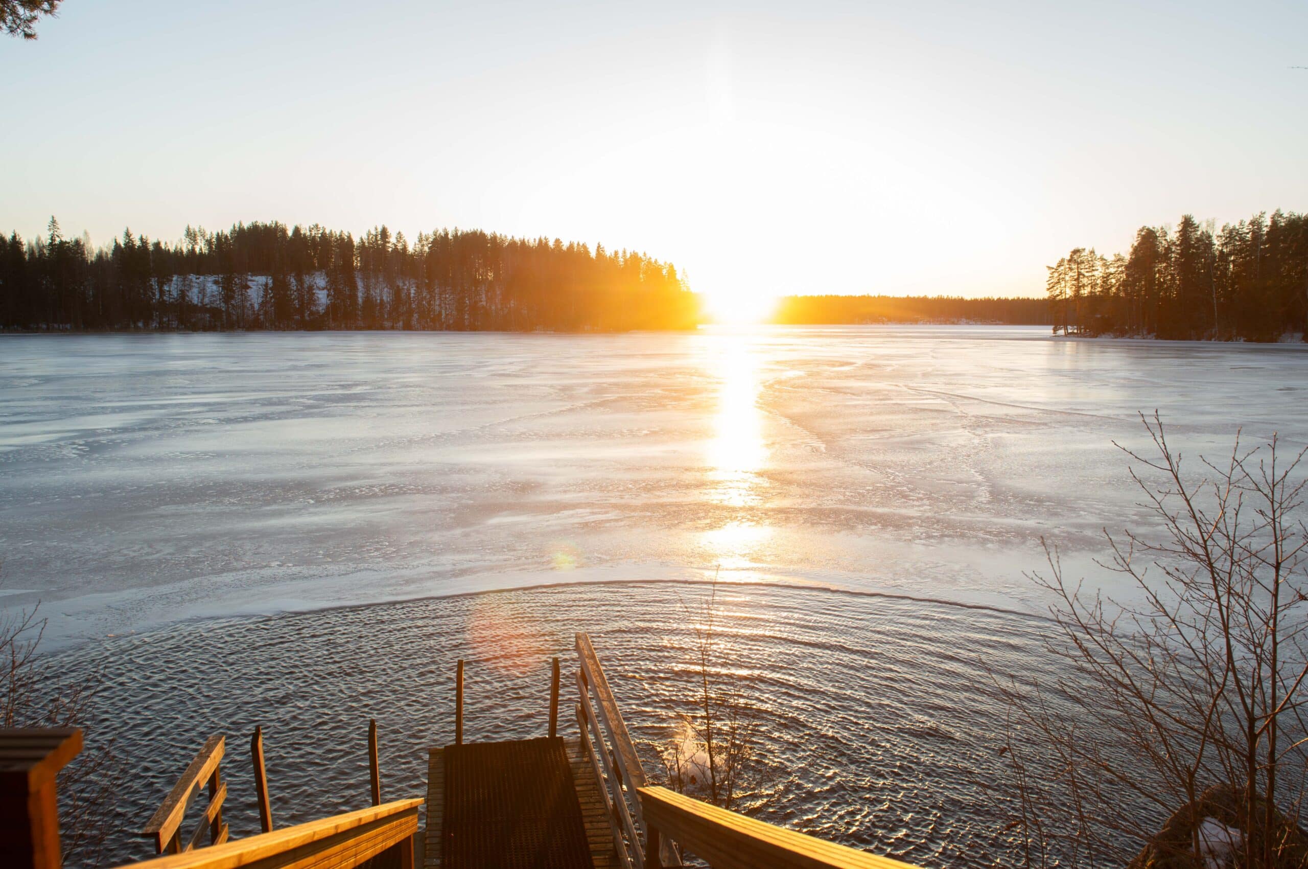 Jäätyneen järven edustalla on sula kohta. Aurinko paistaa puiden takaa.