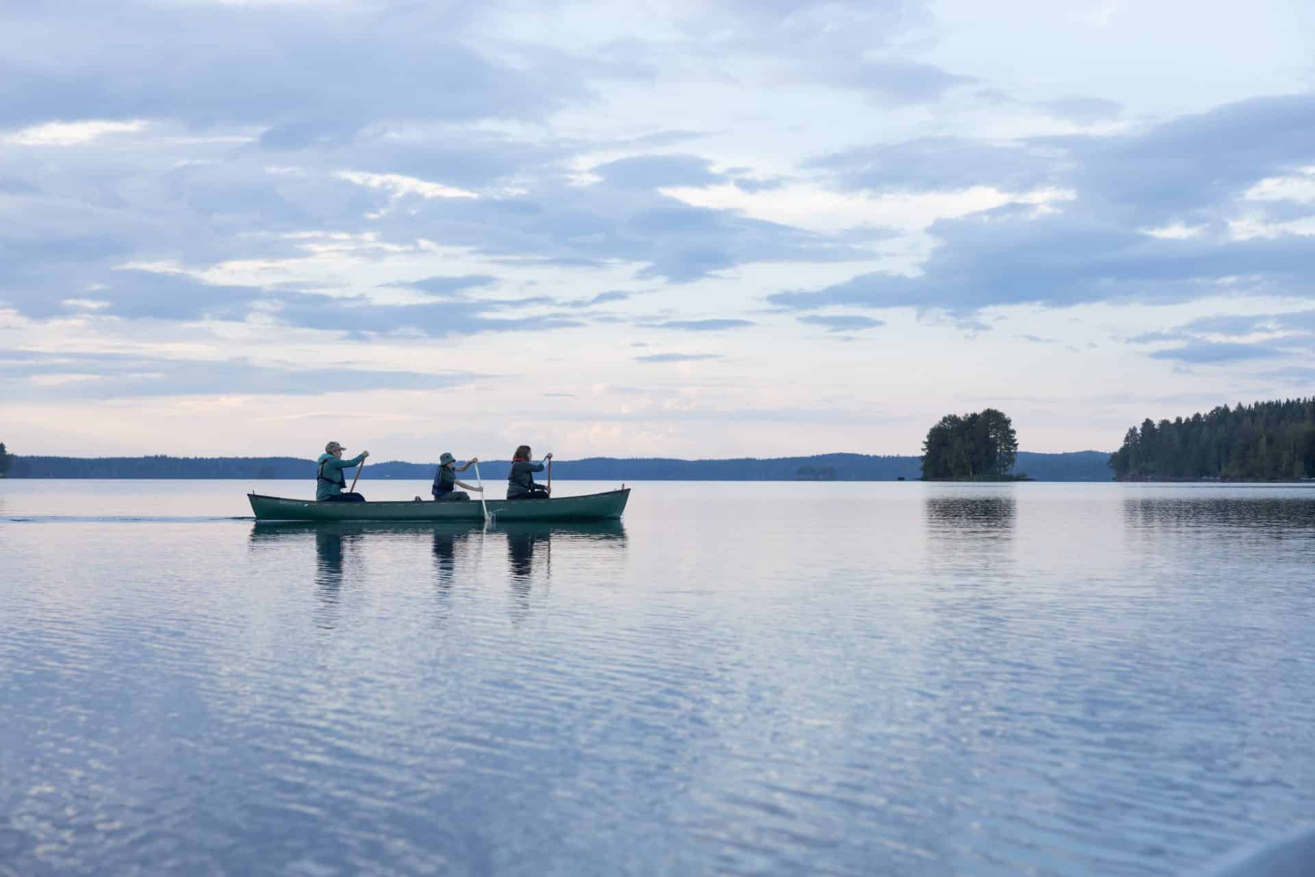 Neljä ihmistä kävelemässä suomaisemassa pitkospuilla, takana korkeita puita, joiden välistä pilkistää auringonsäteitä.