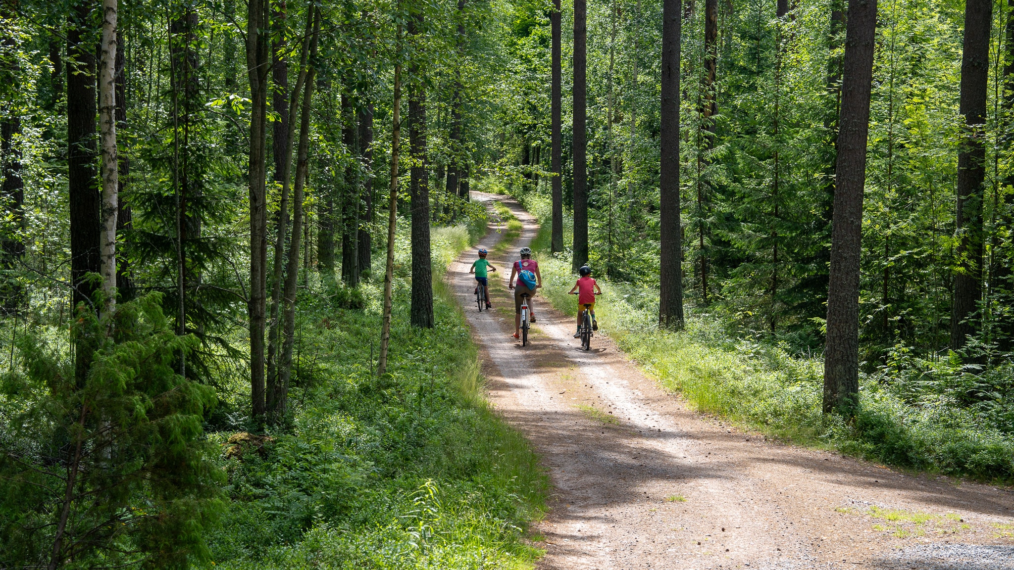 Kolme ihmistä pyöräilevät alamäkeen hiekkatietä pitkin.