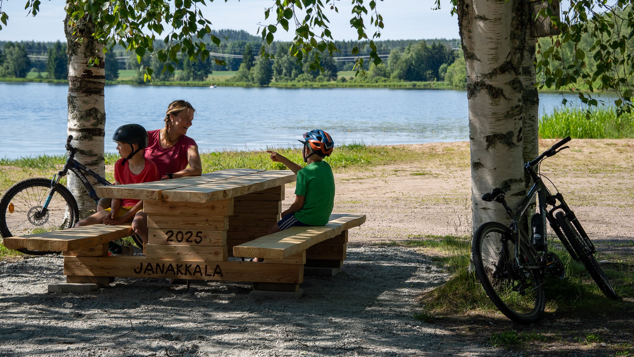 Äiti ja kaksi lasta istuvat puisen pöytäpenkin äärellä järven rannalla.