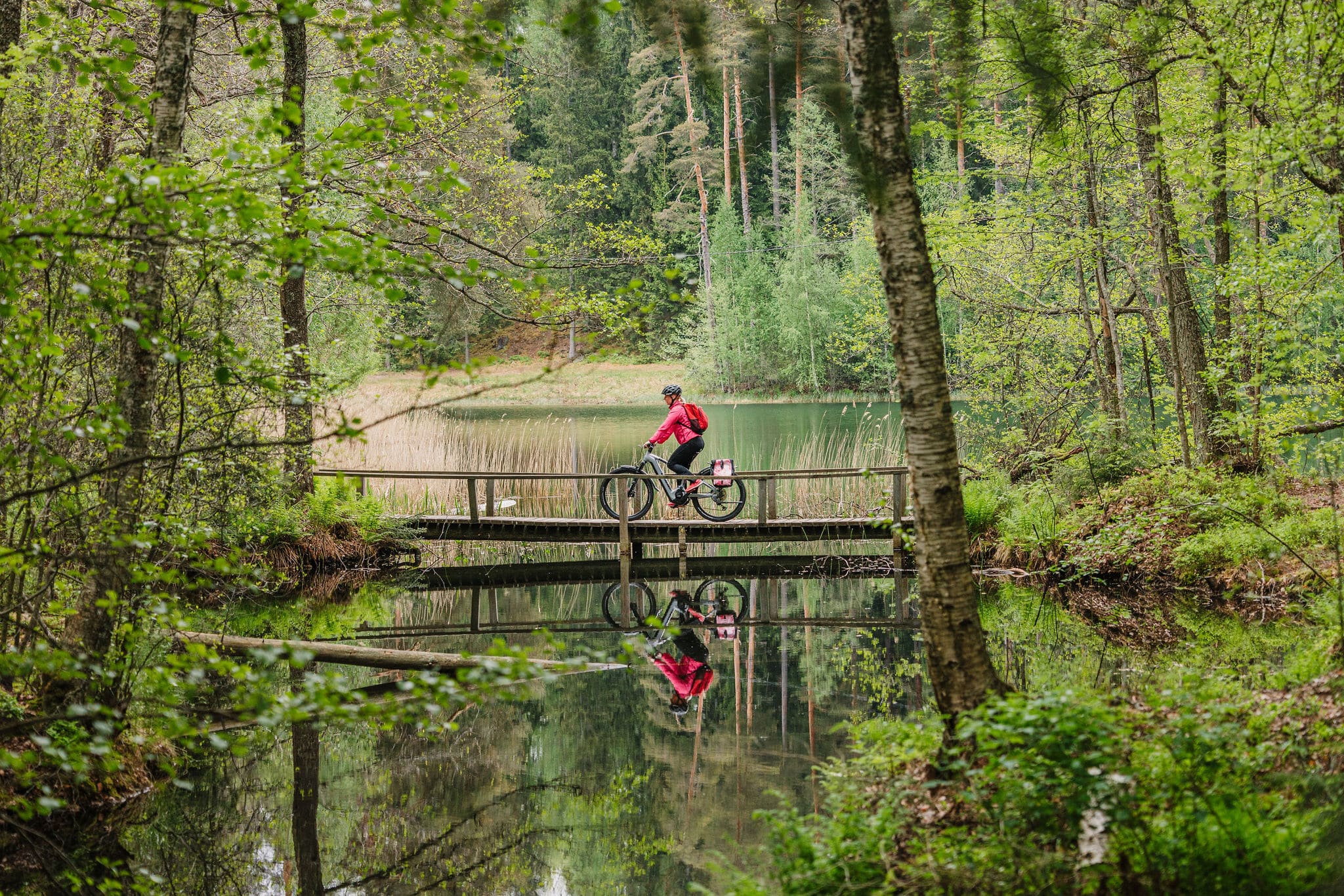 Pyöräilijä on sillalla. Järvestä heijastuu hänen kuvansa.